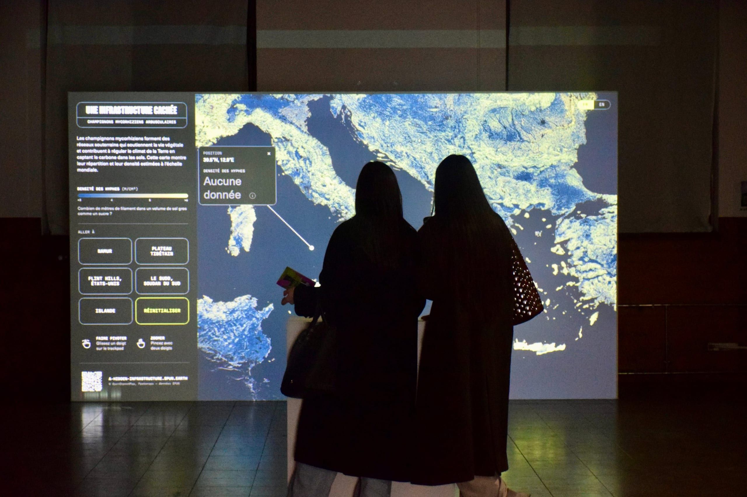 Two visitors in silhouette in front of a large projection of the French-language map, zoomed in on Italy and the Balkans in blue and pale yellow
