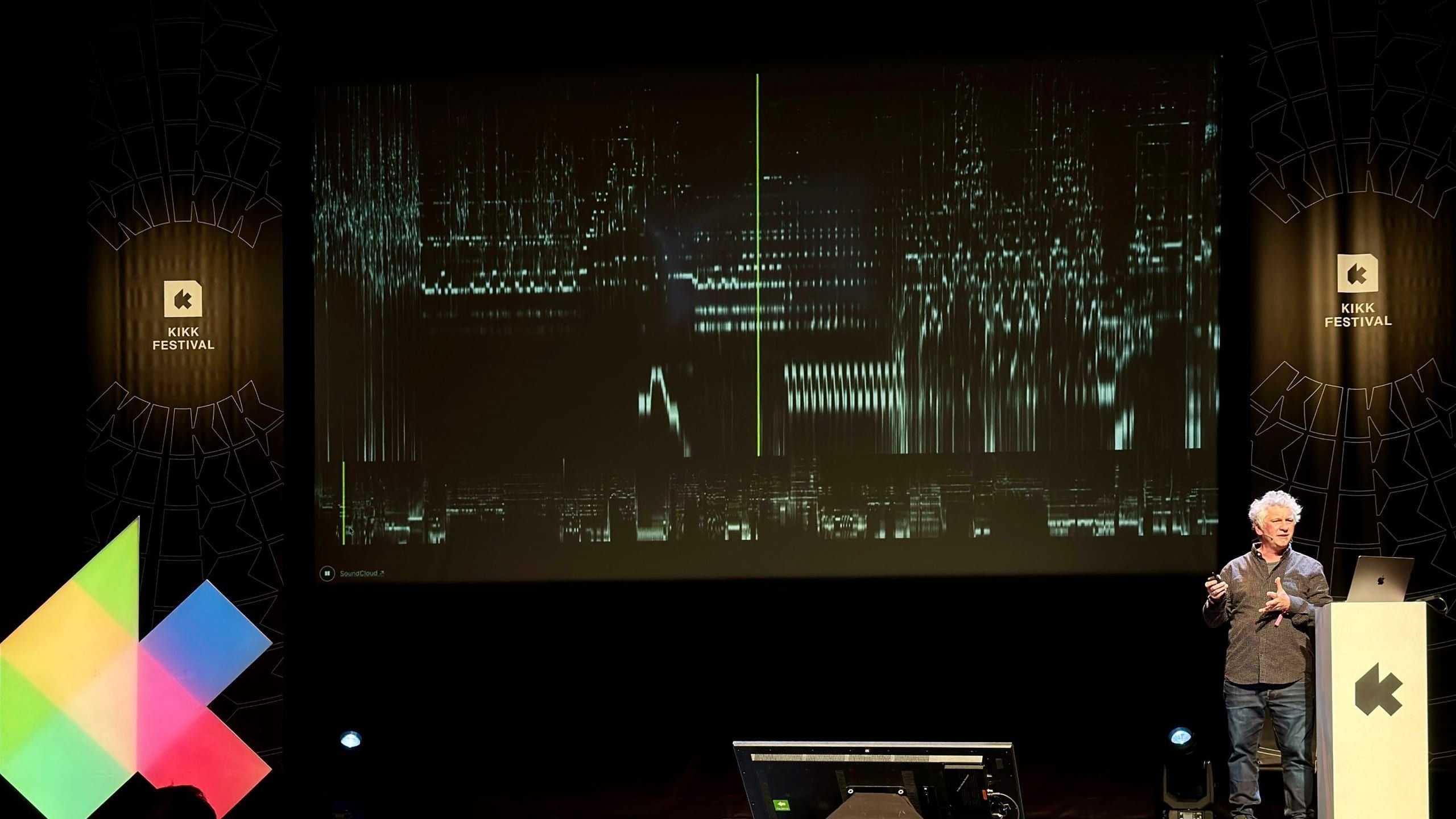 Moritz Stefaner at the KIKK Festival lectern next to a projected spectrogram, bright traces on a black background