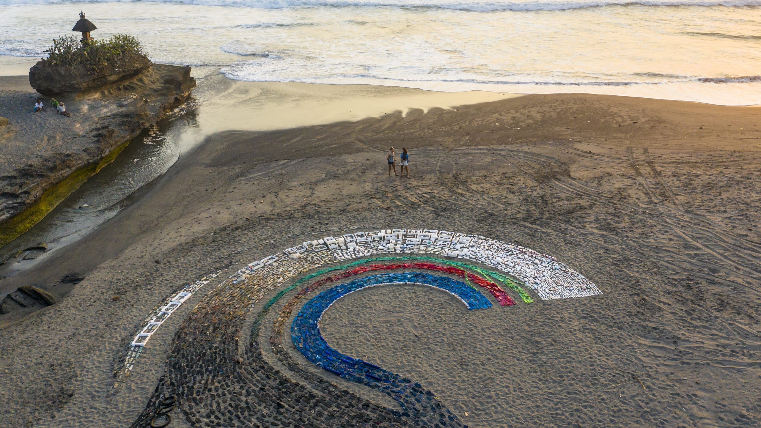 Two people on a Bali beach beside the colorful semi-radial plastic sculpture at sunset, temple shrine on a cliff