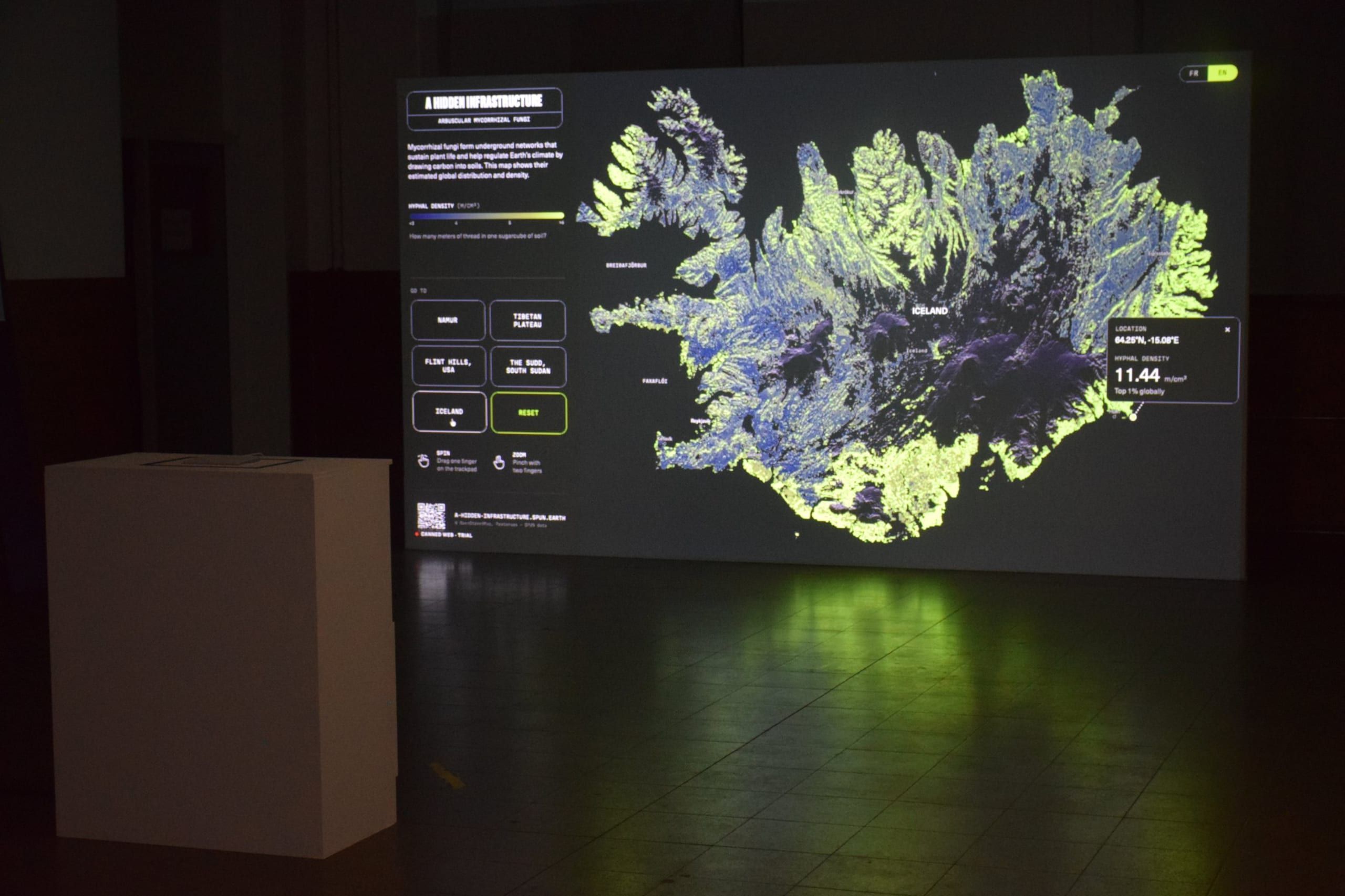 The installation in a darkened hall: a projected map of Iceland in blue and bright green with a tooltip reading 11.44 m/cm³, next to a white plinth holding the trackpad