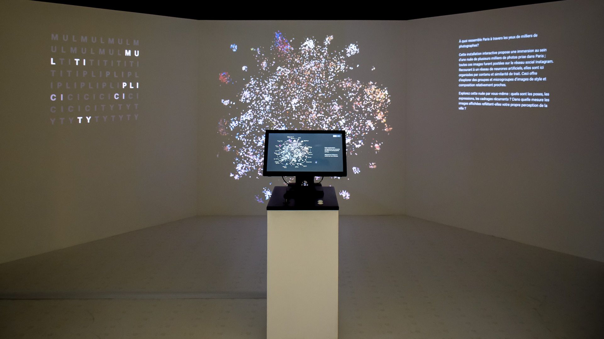 Installation view, touchscreen console before a wide projection of the glowing photo cloud, with title and text on angled walls
