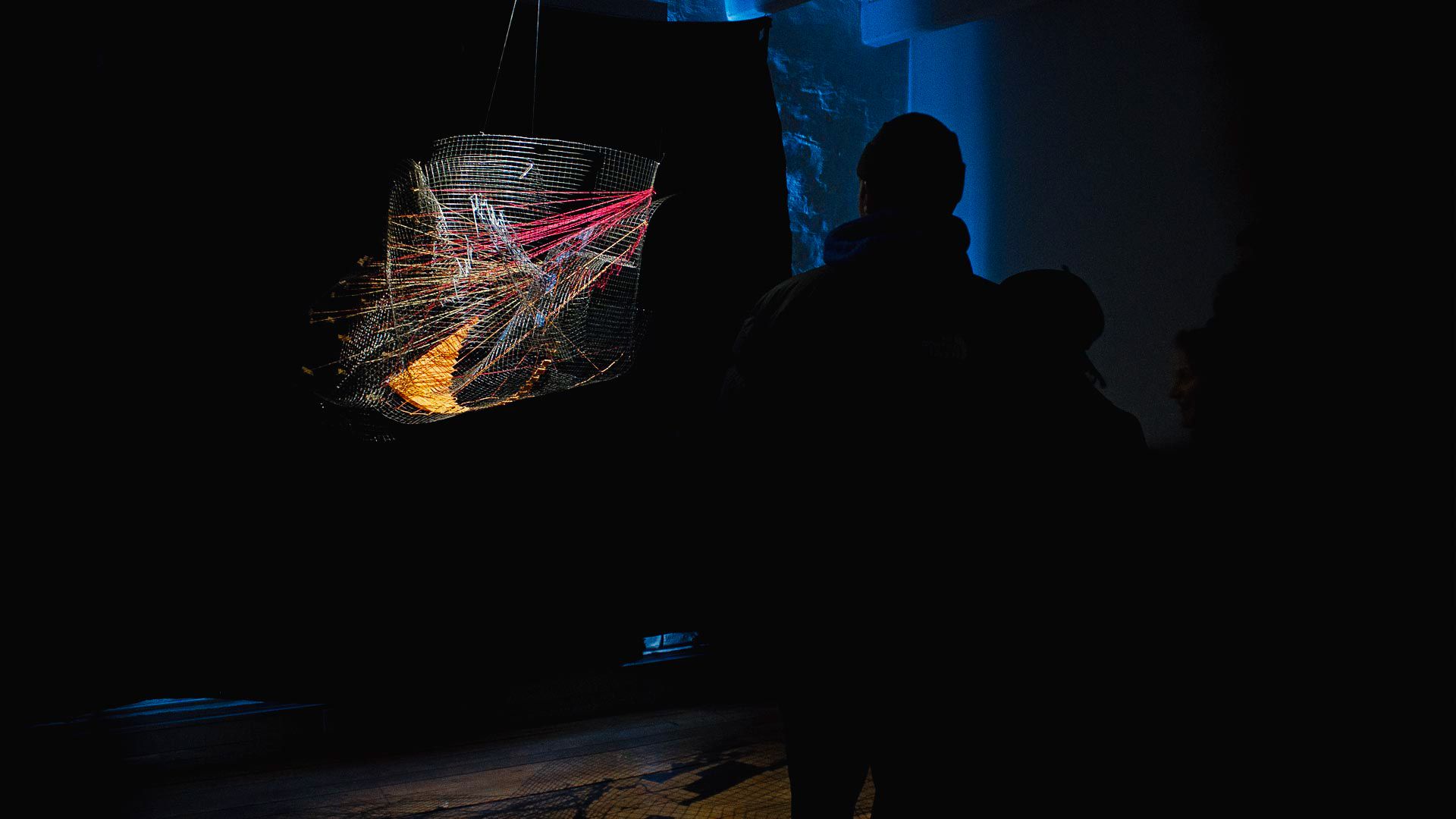 Visitor silhouetted before the glowing HDDN wire-net sculpture in a dark gallery