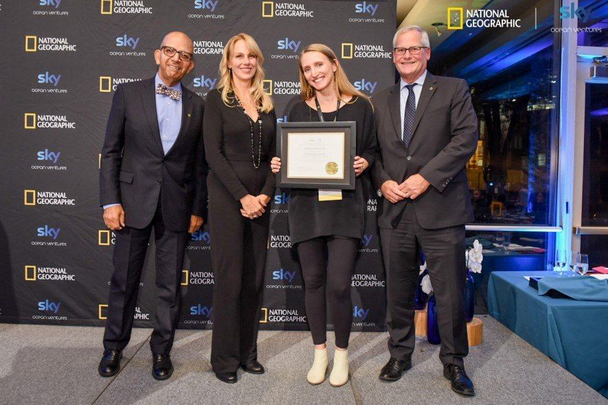 Winner holding the framed award certificate with three presenters at the National Geographic Ocean Plastic Innovation Challenge ceremony