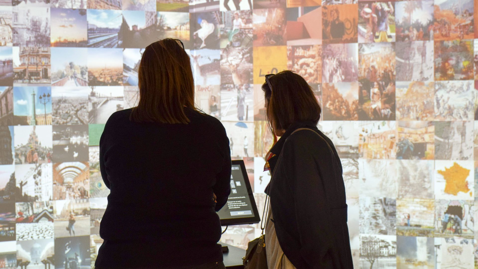 Two visitors at the console before a projected wall of Paris cityscape and landmark photos
