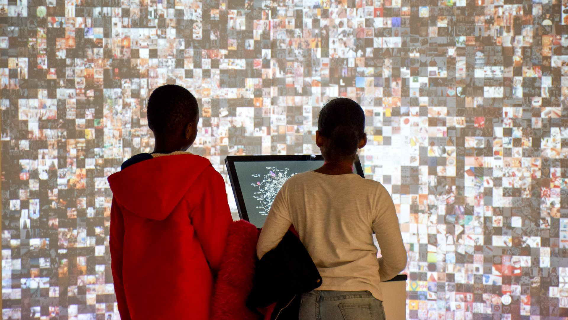 Two visitors seen from behind exploring the map at the console before the full photo-grid projection wall