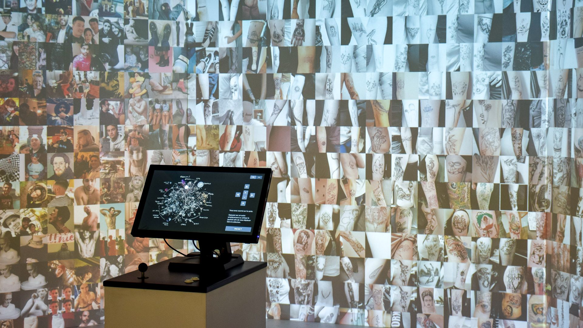 Installation view with joystick console, projected wall zoomed into the tattoos cluster of arms and body art