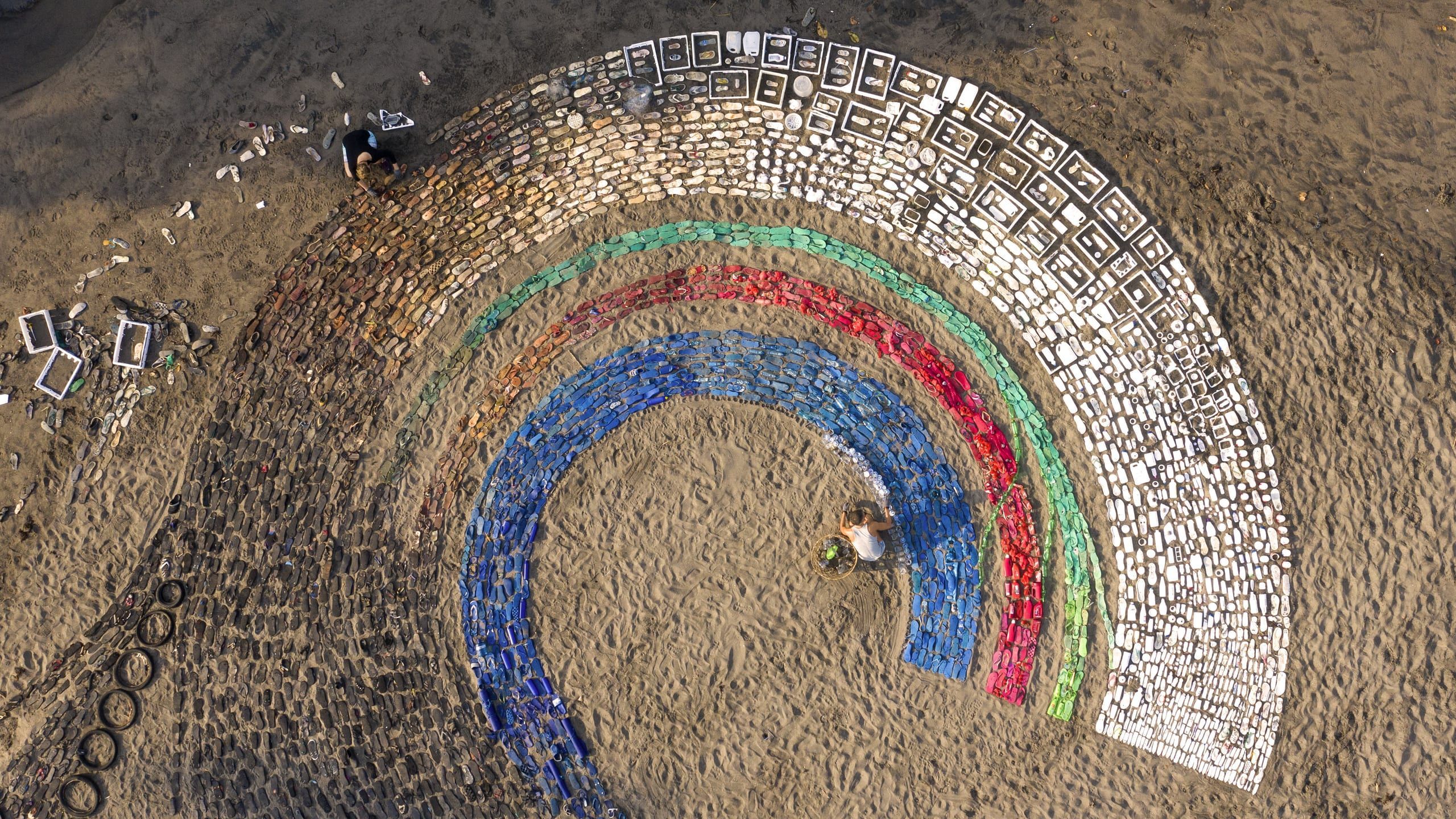 Top-down view of near-complete rainbow arcs of sorted flip-flops and plastic with a volunteer placing pieces